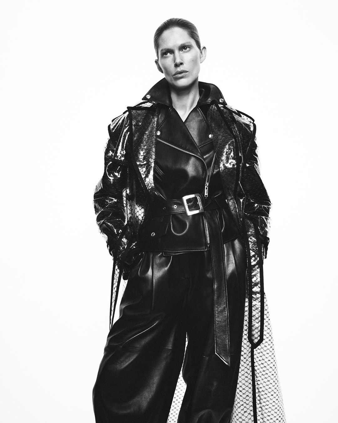 Monochrome fashion shot of an adult female posing in a shiny black belted trench coat and black pants against a plain white backdrop.
