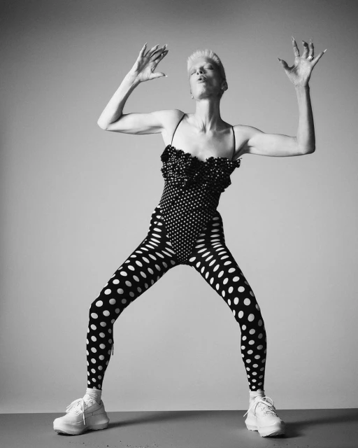 Black and white studio shot of an adult female in a polka-dot outfit, arms raised, against a gradient background. Fashion editorial.