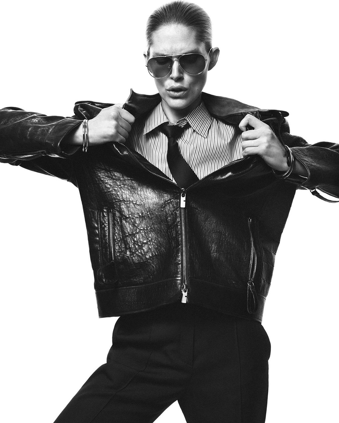 Model in aviator sunglasses wearing a black leather jacket over a striped shirt and tie against a white background.