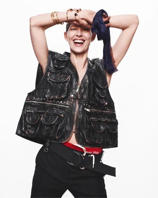 A smiling woman with short hair poses in a black leather vest, black pants, and accessories against a clean white backdrop in a studio shot.