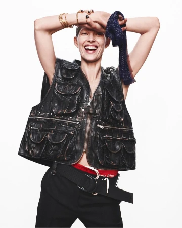 A smiling woman with short hair poses in a black leather vest, black pants, and accessories against a clean white backdrop in a studio shot.