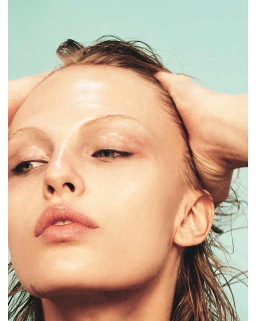 Close-up portrait of a woman with wet hair slicked back, head being held by hands. Light skin tone and a pale blue background.