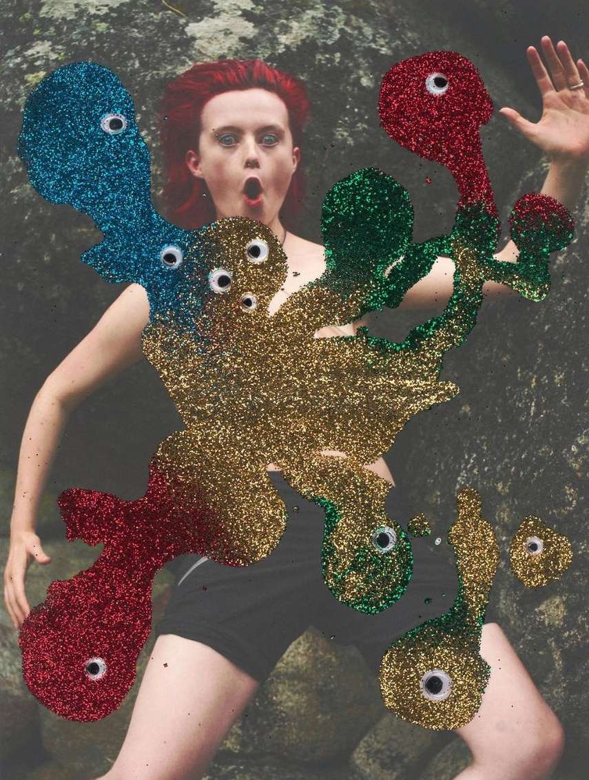 A woman with red hair stands in front of a rock, partially obscured by colorful glitter shapes with googly eyes in a surreal art image.