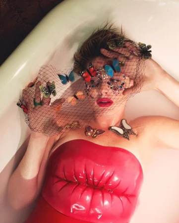 An adult female with an embellished net veil poses in a bathtub, wearing a lip-shaped top and surrounded by faux butterflies in a beauty shot.