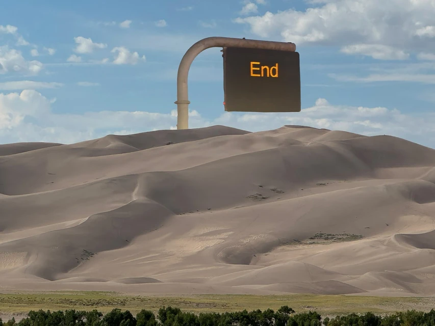 Desert landscape featuring sand dunes beneath a blue sky with white clouds and a sign displaying the word 'End' in orange letters.