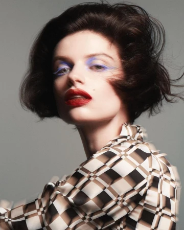 Portrait of a woman with dark bobbed hair wearing blue eyeshadow and red lipstick in a checkered top against a grey background.