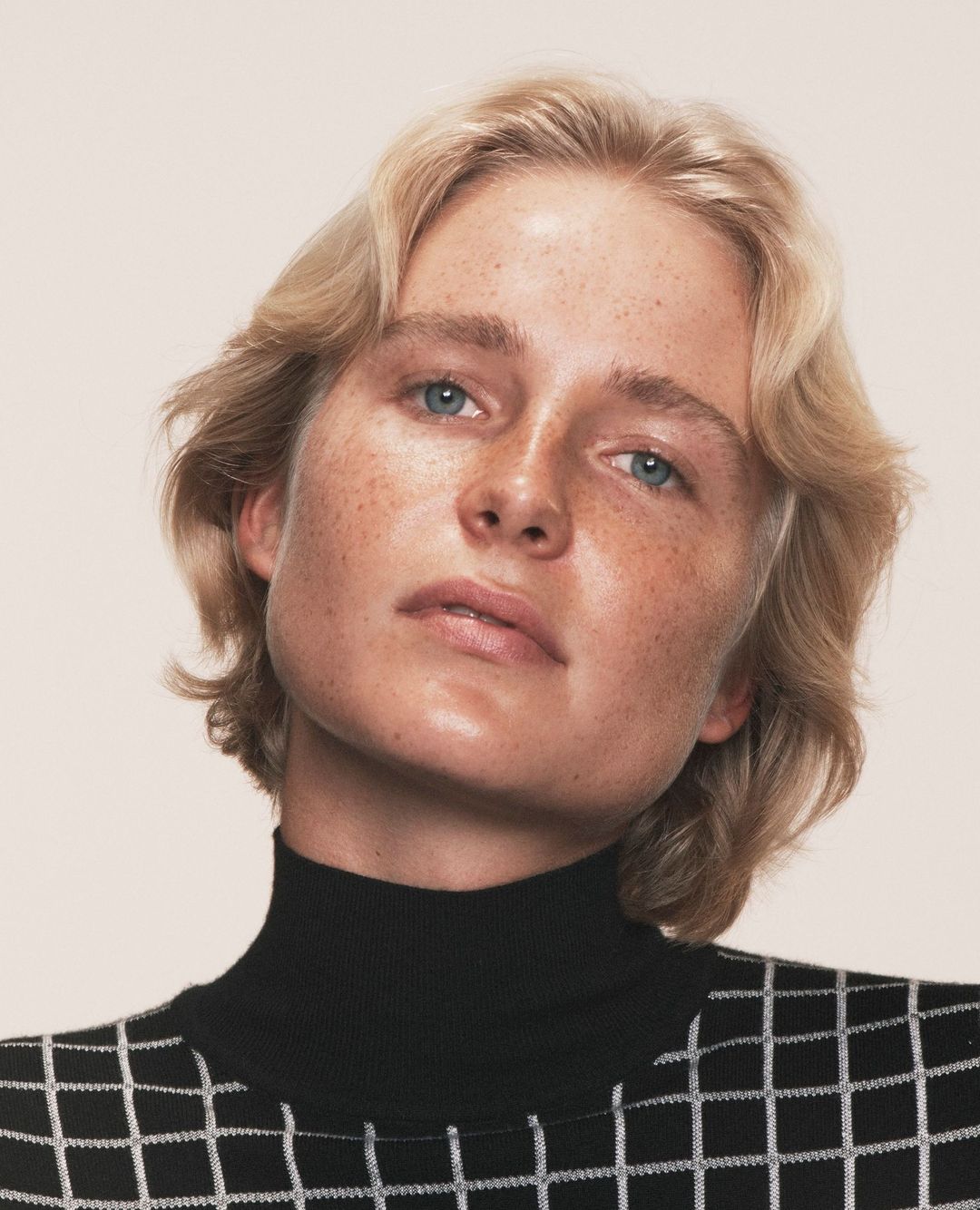 Portrait of a blonde woman with freckles wearing a black turtleneck with white grid pattern against a light background