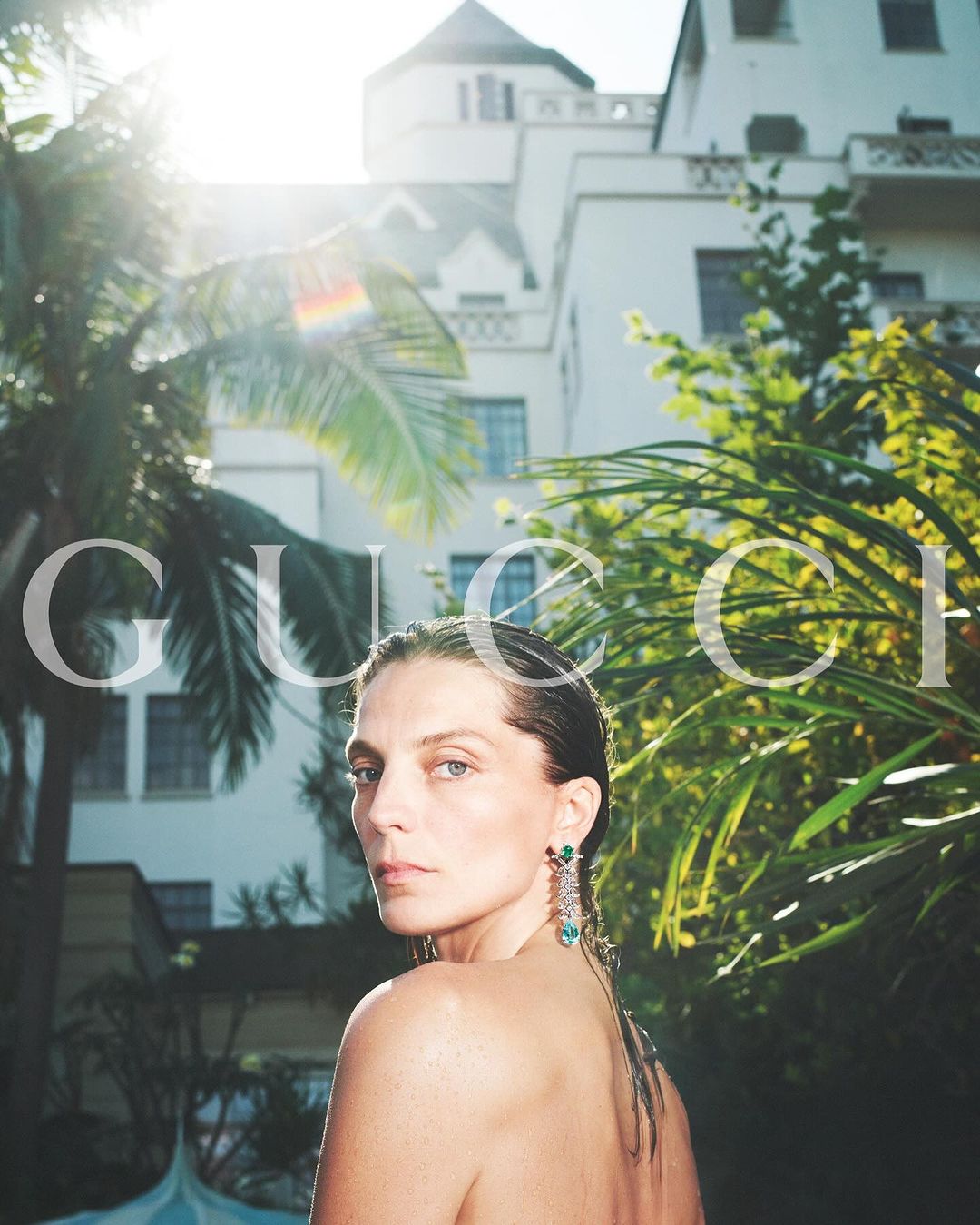 Fashion editorial shot of an adult woman with wet hair and dangling earrings, posing in a lush garden in front of a building.