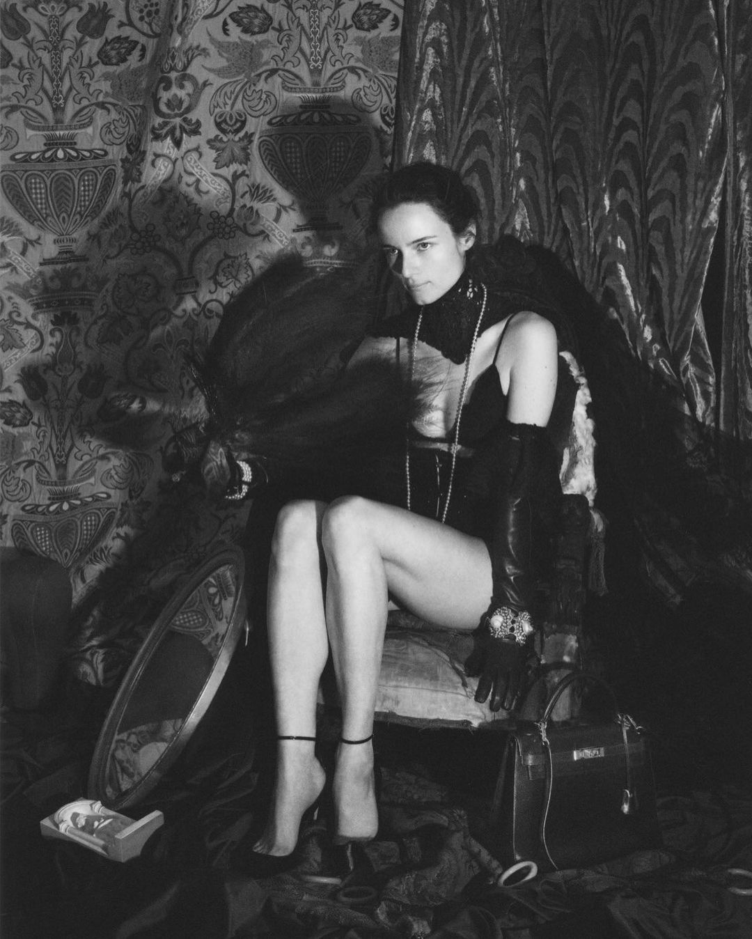 A black and white portrait of a Caucasian woman sitting in a chair with an elegant choker, pearls, gloves, dress, and purse.