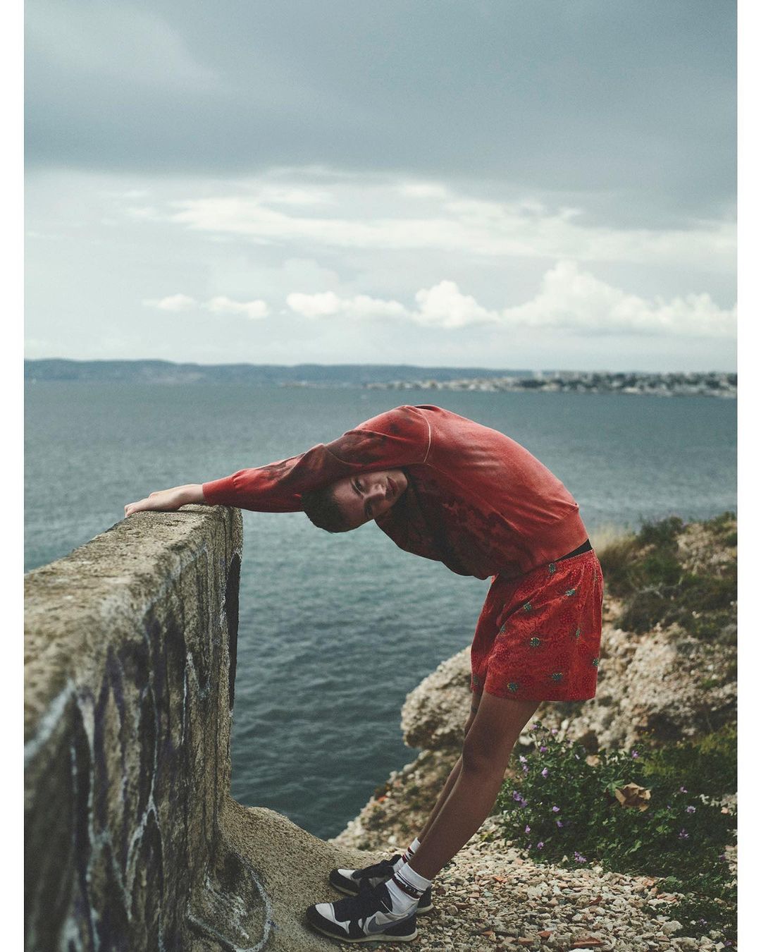 Adult female in sportswear arches back on a concrete wall by the sea on an overcast day. Visible are the ocean, a cloudy sky and distant land.