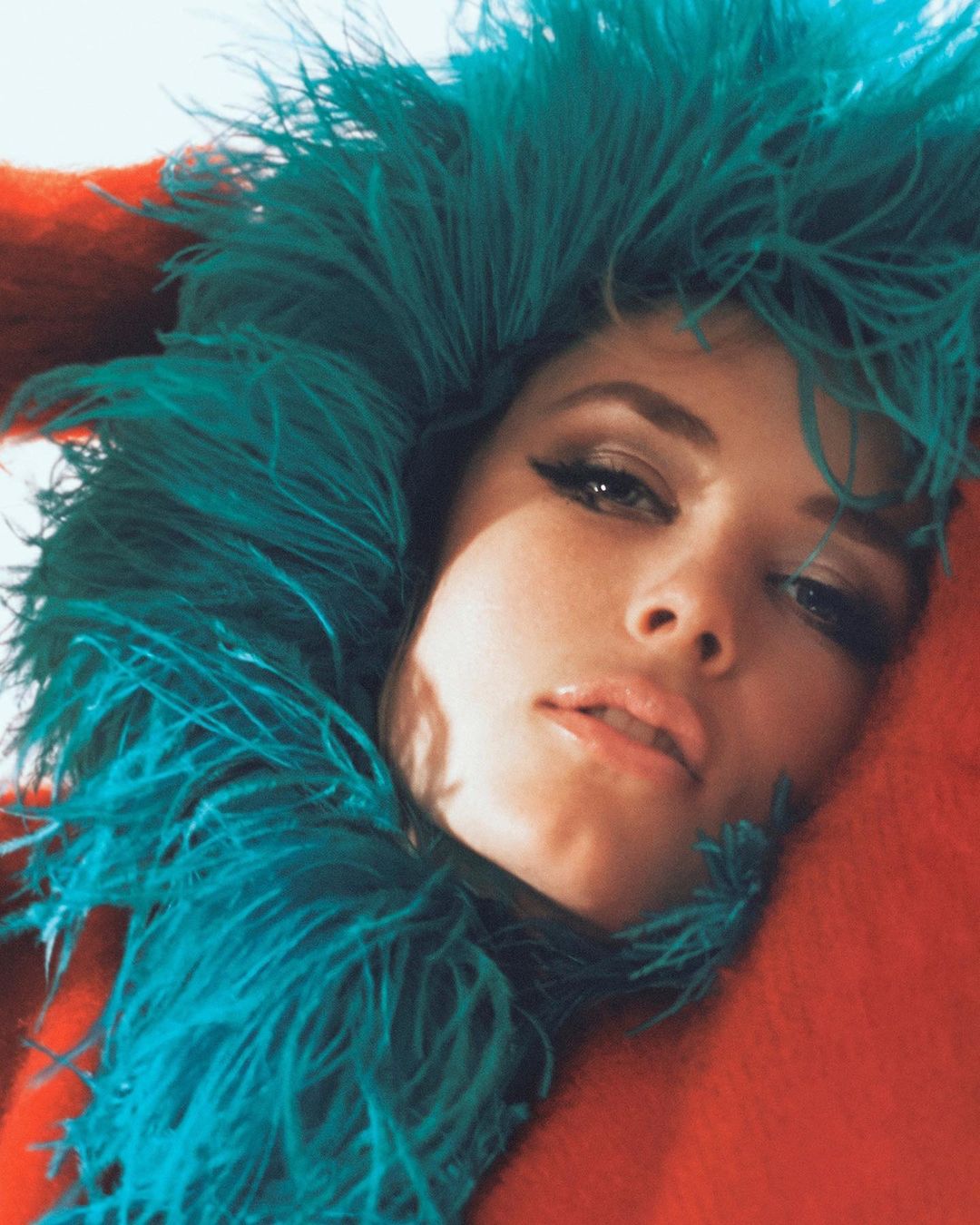 Close-up portrait of a woman wearing a vibrant teal feathered hood against a warm red textured background.