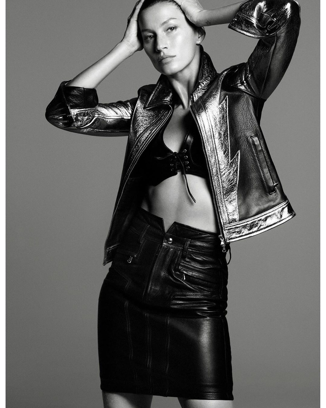 Monochrome fashion shot: woman in a metallic jacket and leather mini skirt poses with hands on her head in front of gray backdrop.