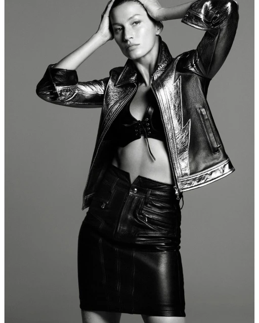 Monochrome fashion shot: woman in a metallic jacket and leather mini skirt poses with hands on her head in front of gray backdrop.