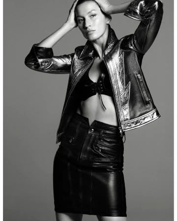 Monochrome fashion shot: woman in a metallic jacket and leather mini skirt poses with hands on her head in front of gray backdrop.