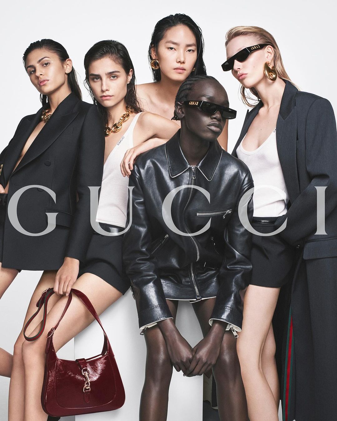 Group of four female models posing in dark jackets and white tops against a white background with prominent Gucci text.