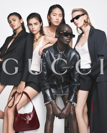 Group of four female models posing in dark jackets and white tops against a white background with prominent Gucci text.