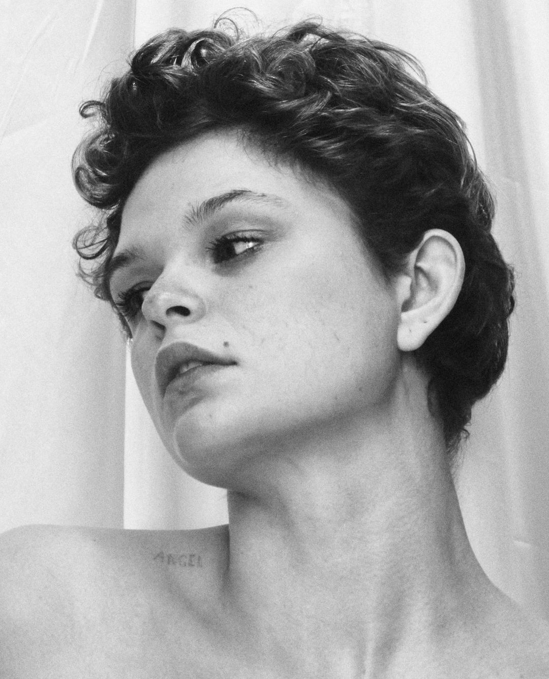 Monochrome portrait of a woman with short curly hair and a shoulder tattoo in a clean, white studio setting, capturing a minimalist aesthetic.