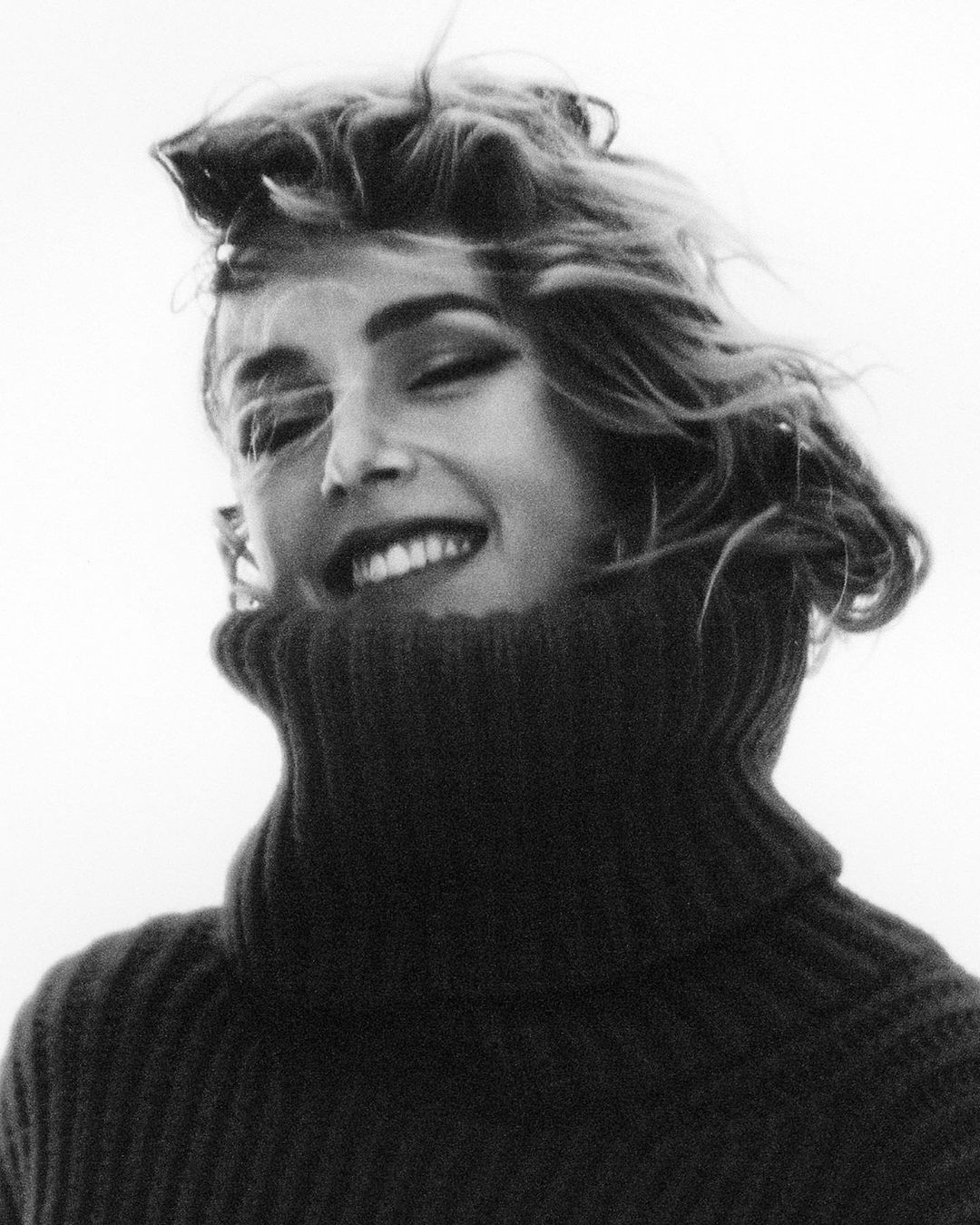 Monochromatic portrait of a young adult female smiling, wearing a dark sweater with a high turtleneck, and a bright white background.