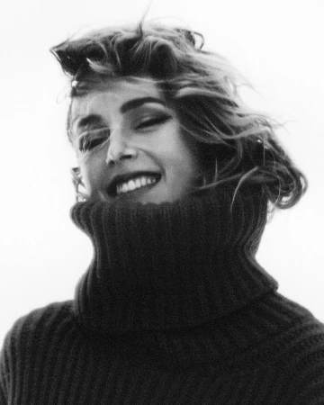Monochromatic portrait of a young adult female smiling, wearing a dark sweater with a high turtleneck, and a bright white background.