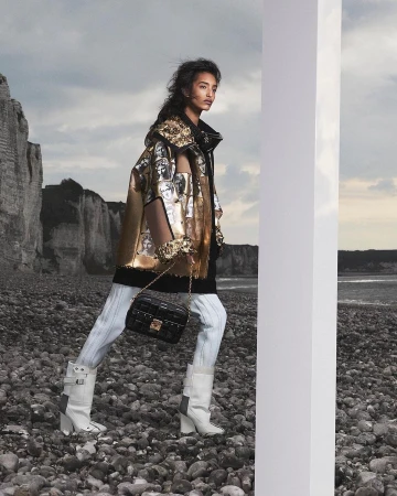 Model in metallic gold printed jacket holding a black quilted handbag walking on a pebbled beach