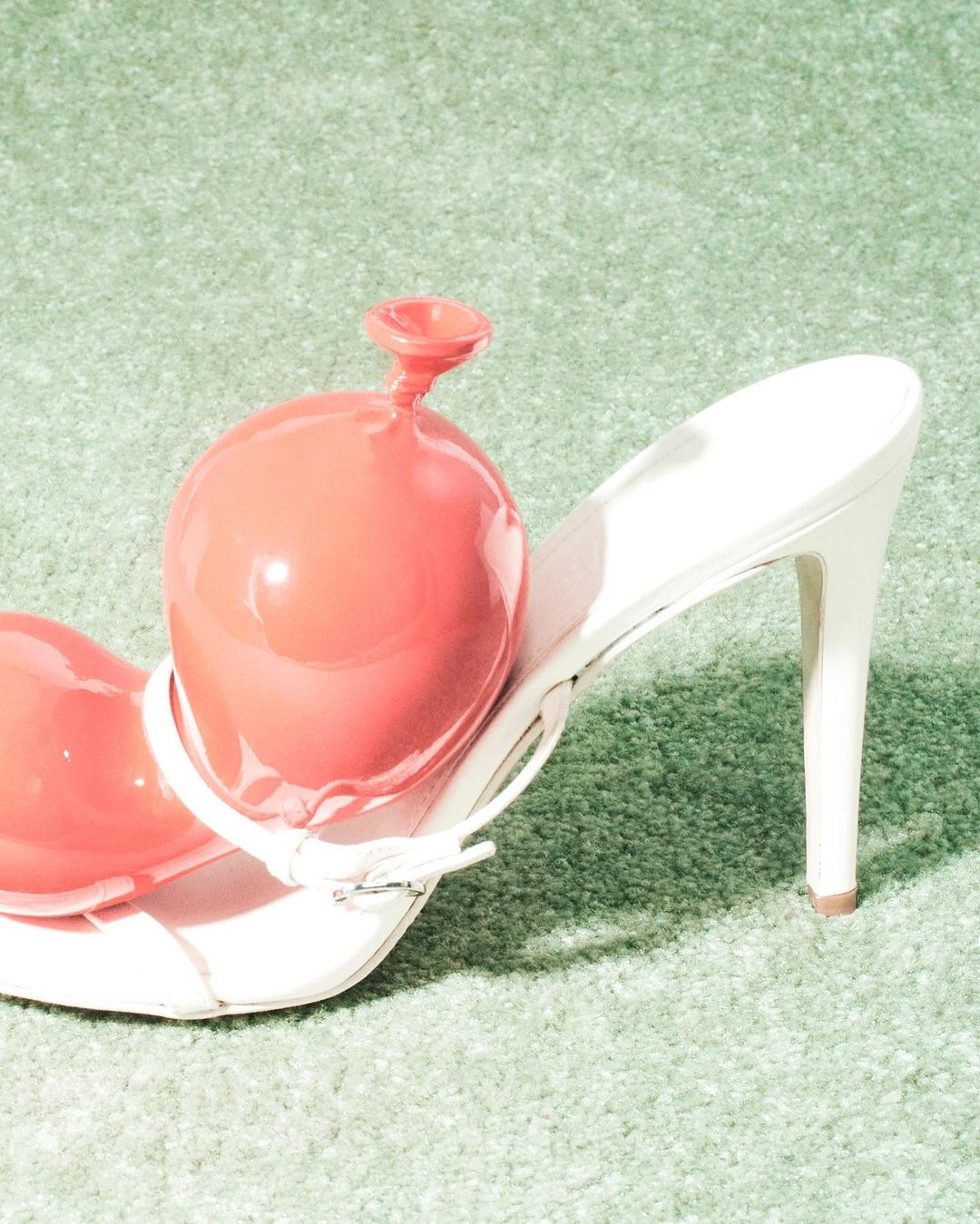 Fashion still life: A salmon-colored water balloon rests inside a white high-heeled mule shoe against a soft, light green carpet backdrop.