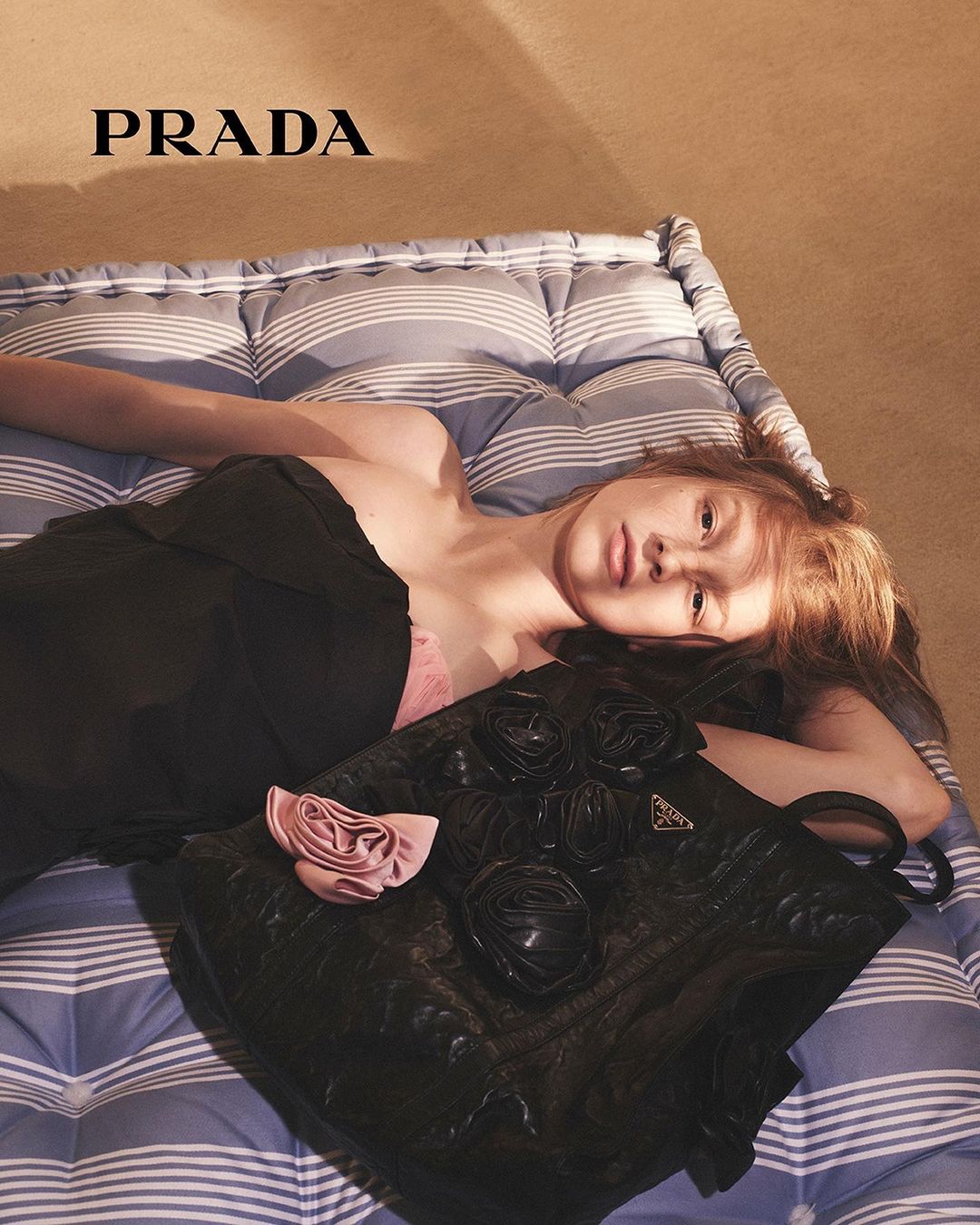 A woman lying on a striped mattress next to a black floral handbag with PRADA text visible in the upper corner.