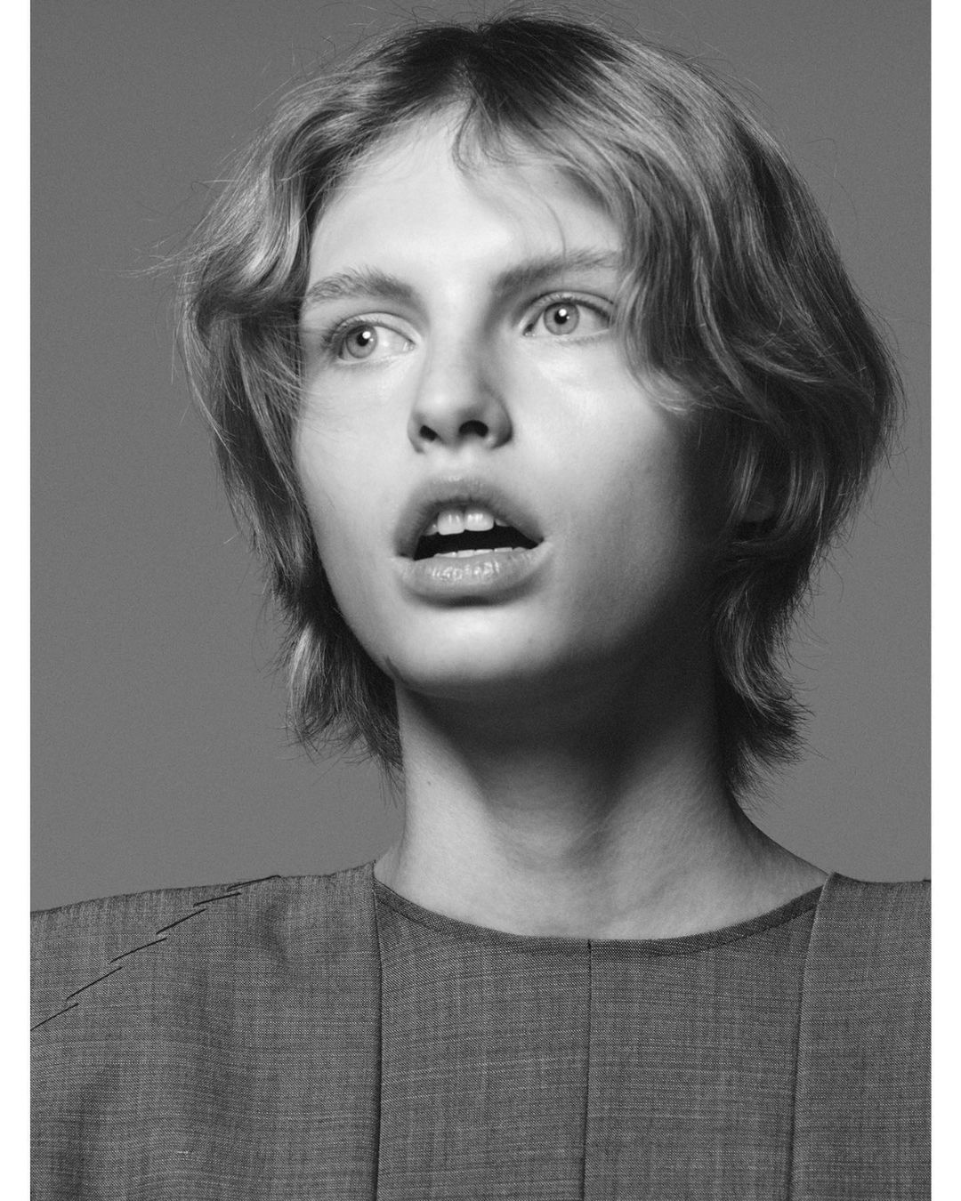 Monochrome close-up studio portrait of a fair-skinned young adult with wavy blonde hair wearing a gray crew neck top looking right.