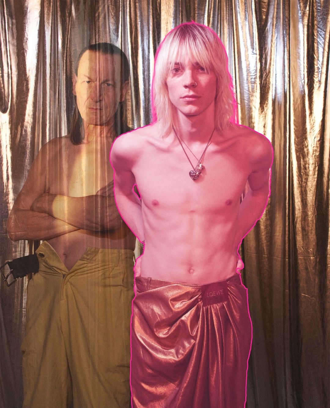 A young man with long blonde hair stands in front of a shiny gold curtain with a semi-transparent man in the background.