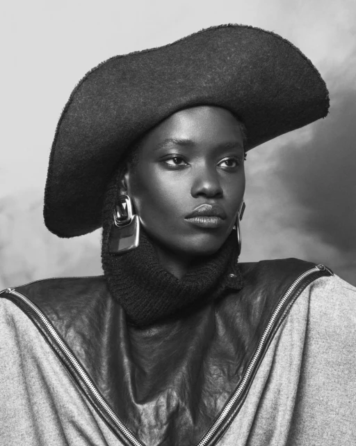 Monochrome portrait of a Black woman wearing a wide-brimmed hat, chunky earrings, a turtleneck, and a zipper-trimmed top against a cloudy backdrop.