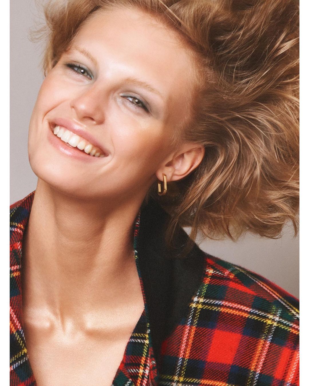 Close-up of a woman with short red hair, smiling, wearing gold earrings and a red and green plaid jacket against a grey background.