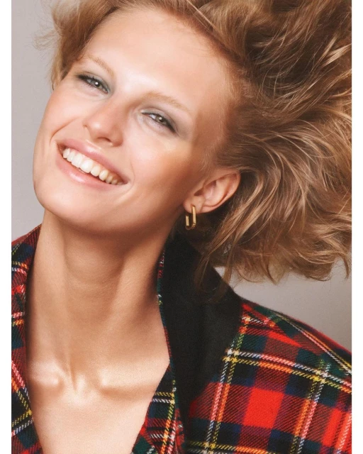 Close-up of a woman with short red hair, smiling, wearing gold earrings and a red and green plaid jacket against a grey background.