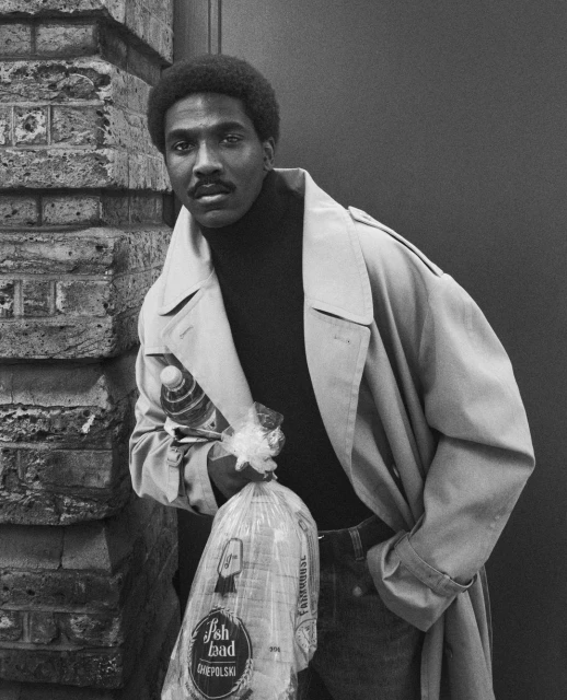 Monochromatic portrait of African-American man in trench coat with baguettes and a water bottle next to a brick wall.