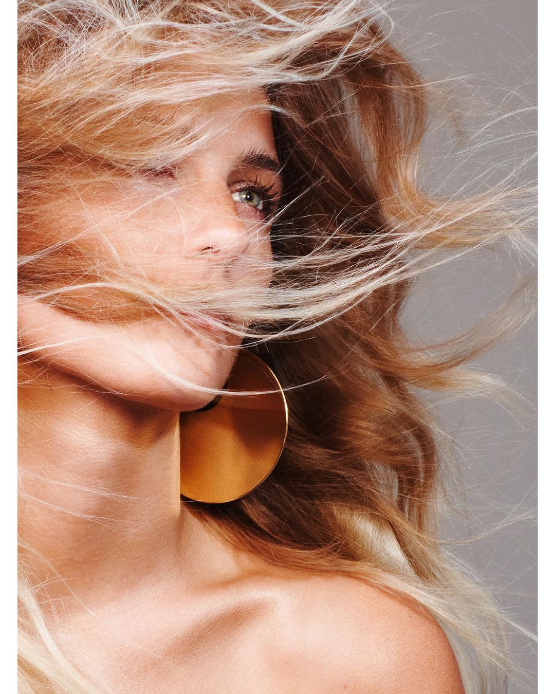 Close-up portrait of a blonde woman with statement gold hoop earrings, windswept hair, and shallow depth of field against a grey background.