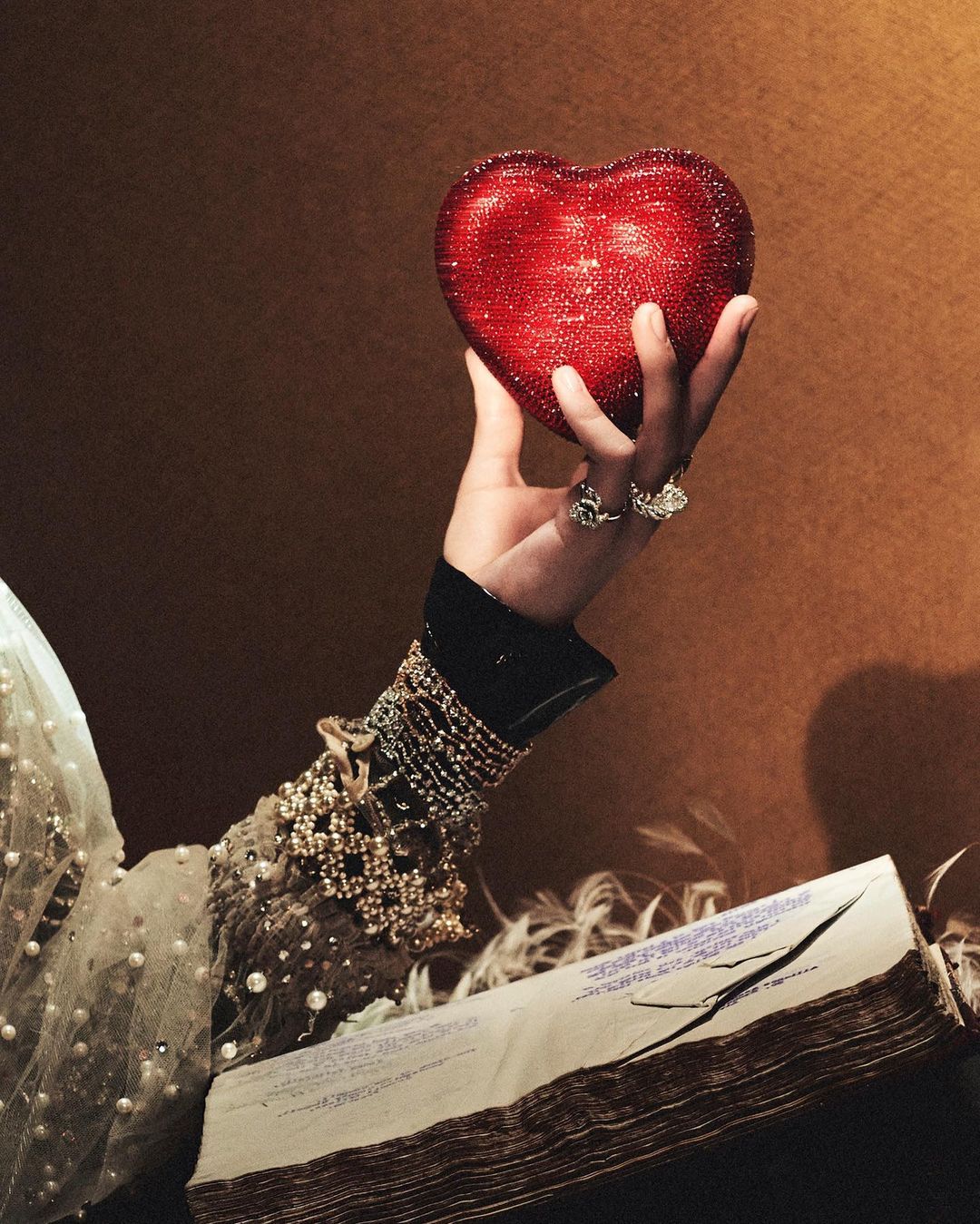 Close-up of a woman's hand holding a red, heart-shaped object with an open book in the foreground. Gold and brown background.