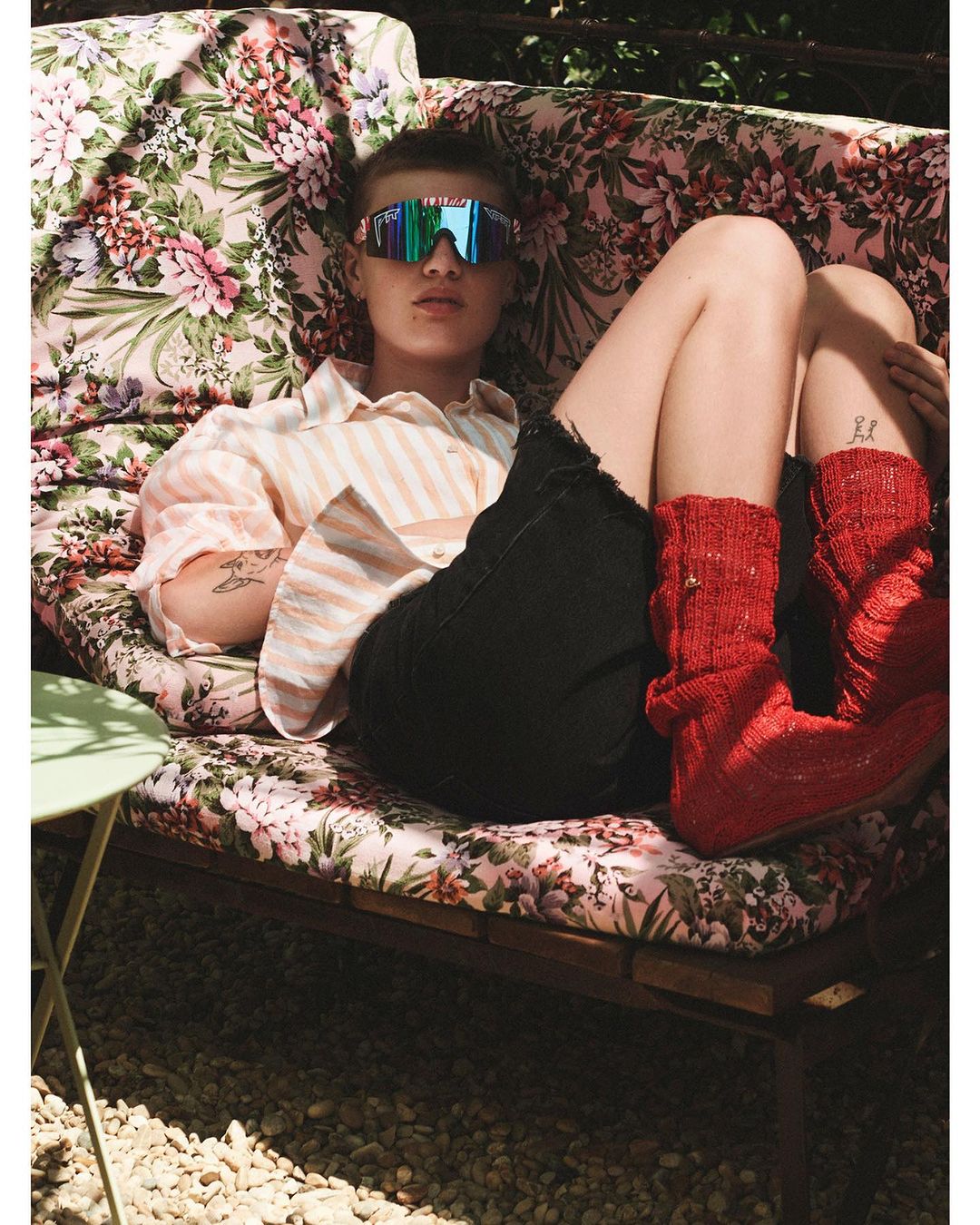 Model wearing mirrored sunglasses, striped shirt, and bright red socks lounging on a floral patterned couch outdoors.