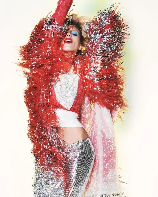 A woman wearing a sparkling silver outfit and a red feathered jacket poses with her arms raised in a fashion editorial shot.