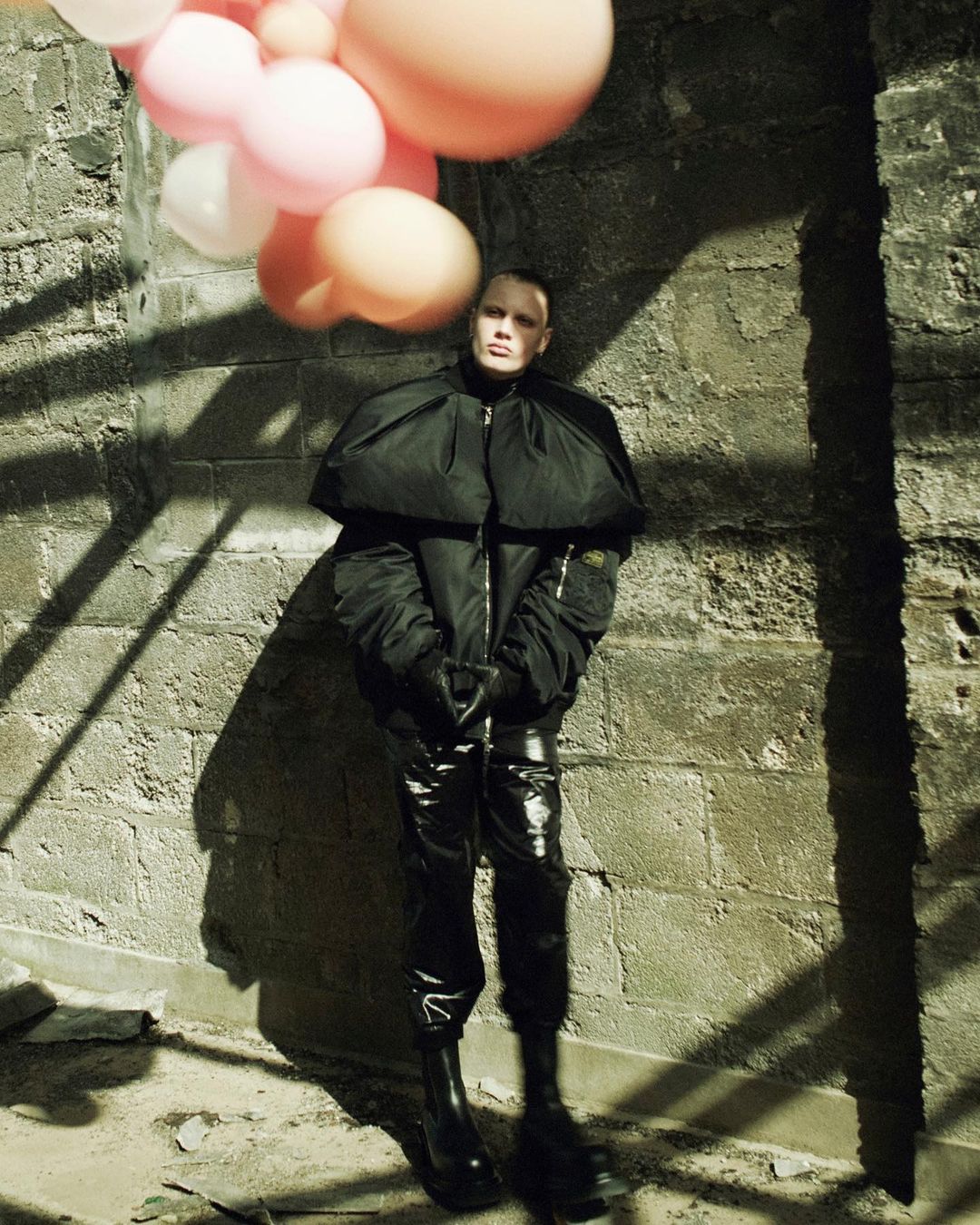 Person poses in black jacket and vinyl pants next to brick wall, holding balloons in shadow-casted light, fashion and editorial.