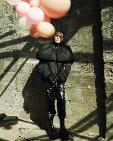 Person poses in black jacket and vinyl pants next to brick wall, holding balloons in shadow-casted light, fashion and editorial.