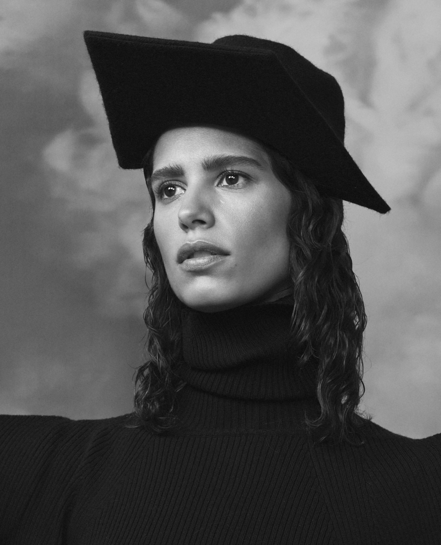 Monochrome portrait of a woman with dark wavy hair wearing a black turtleneck and a square-shaped hat against a soft cloud background.
