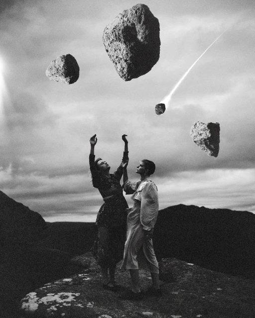 Two women stand on a rocky surface with floating asteroids above them, set against a cloudy sky in a black and white outdoor scene.