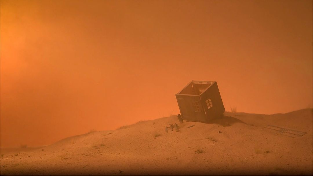 Dusty landscape with a small, derelict structure leaning precariously on a sandy hill under a hazy, orange sky. Post-apocalyptic scene.