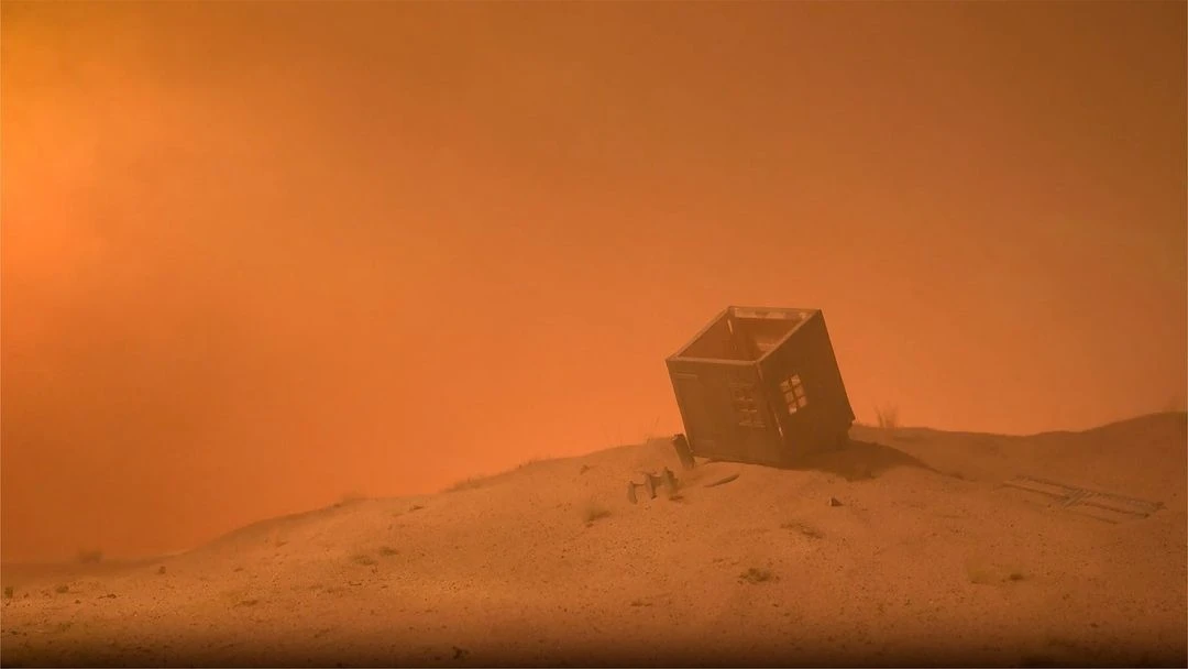 Dusty landscape with a small, derelict structure leaning precariously on a sandy hill under a hazy, orange sky. Post-apocalyptic scene.