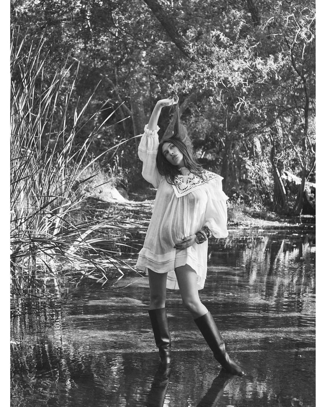 Woman in white dress and boots stands in shallow water in a sun-dappled, monochrome forest scene. Fashion editorial, nature setting.