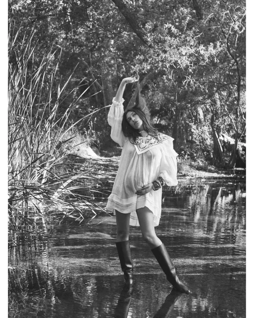 Woman in white dress and boots stands in shallow water in a sun-dappled, monochrome forest scene. Fashion editorial, nature setting.