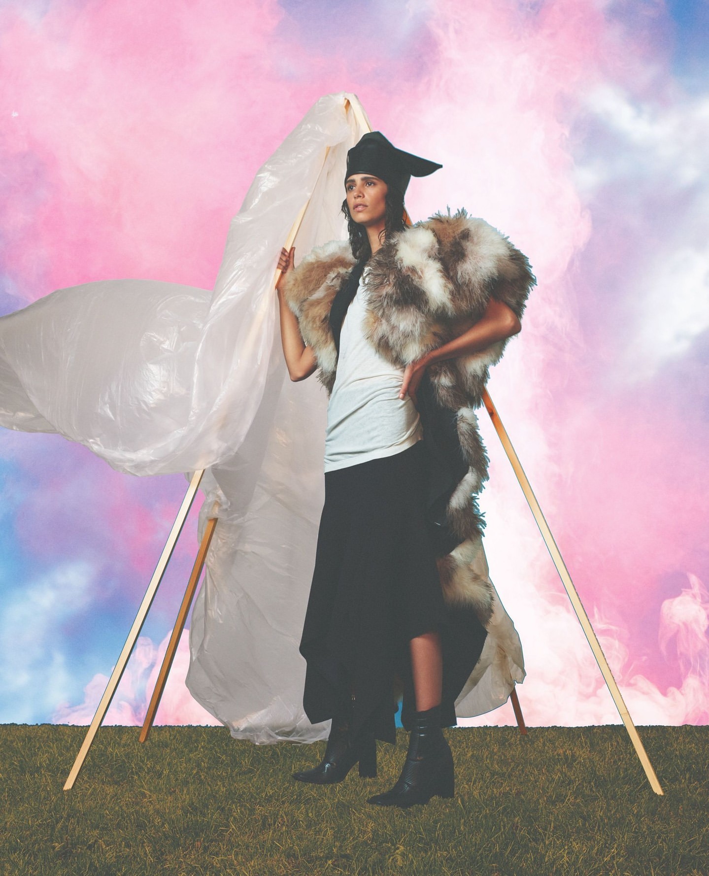 A woman in a fur-trimmed outfit poses outdoors with an abstract prop and a pink, cloudy sky background, captured in a fashion-style photograph.