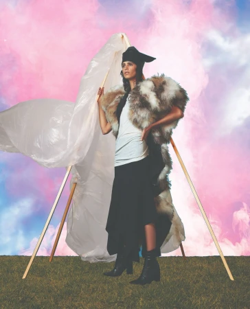 A woman in a fur-trimmed outfit poses outdoors with an abstract prop and a pink, cloudy sky background, captured in a fashion-style photograph.