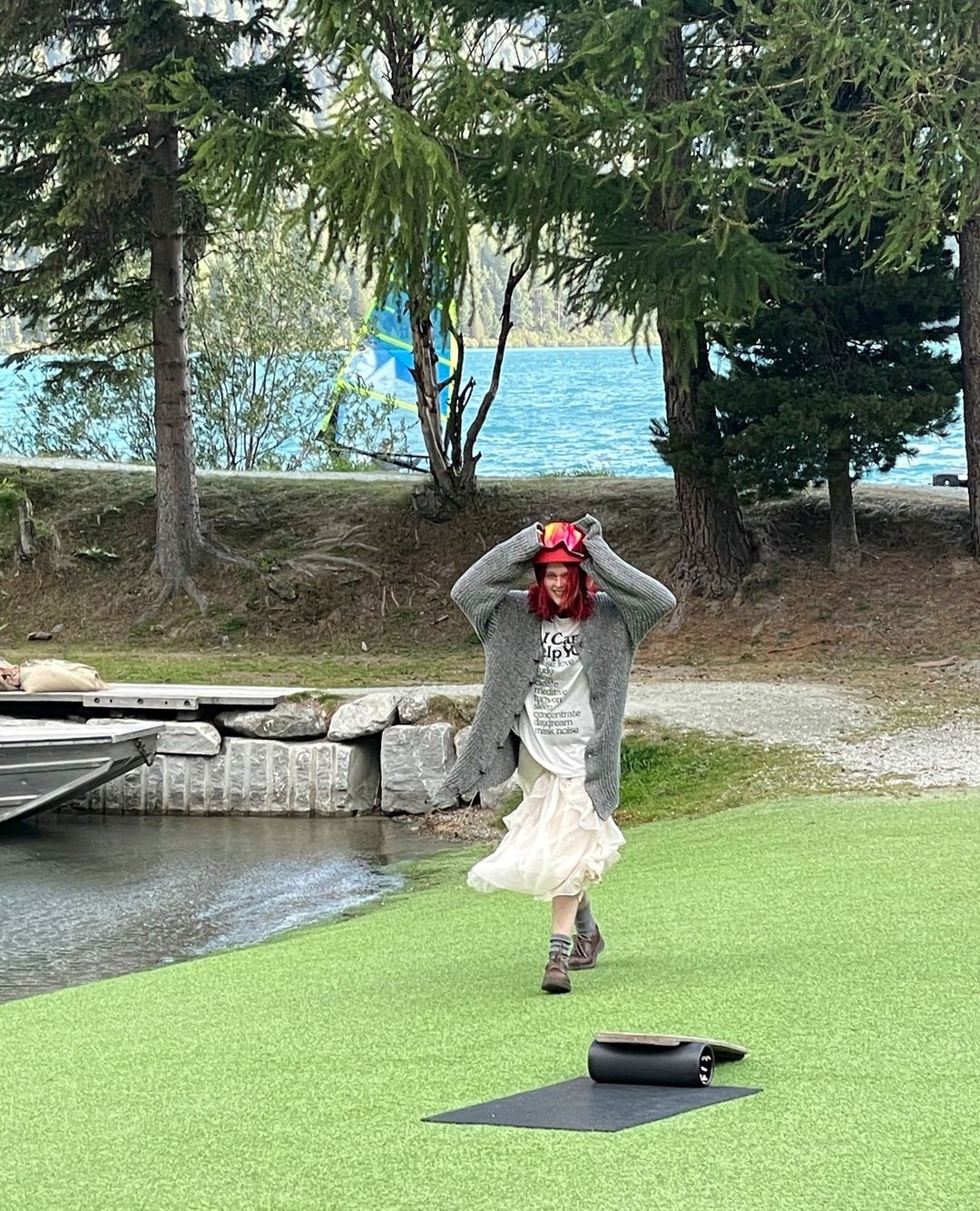 Adult woman walking on green artificial turf near lake and trees wearing ski goggles on head, gray cardigan, boots and a white dress.