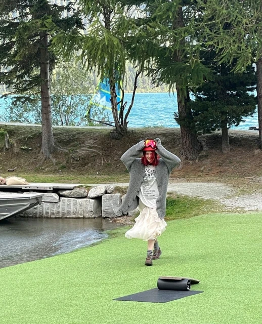Adult woman walking on green artificial turf near lake and trees wearing ski goggles on head, gray cardigan, boots and a white dress.