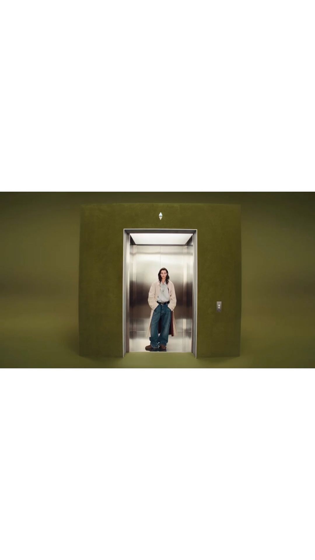 An adult stands in an elevator with an olive green exterior, wearing a pale jacket and jeans, against a solid olive green backdrop.