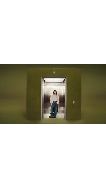 An adult stands in an elevator with an olive green exterior, wearing a pale jacket and jeans, against a solid olive green backdrop.